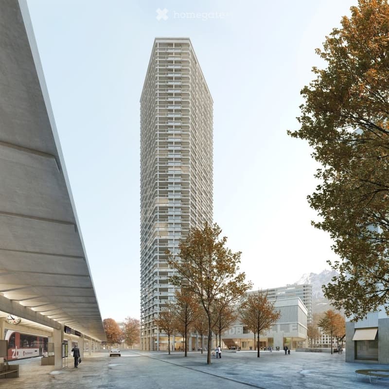A modern, tall residential building with a grid-like facade stands prominently in an urban setting, surrounded by trees and a spacious plaza with a few pedestrians and a tram station visible in the background.