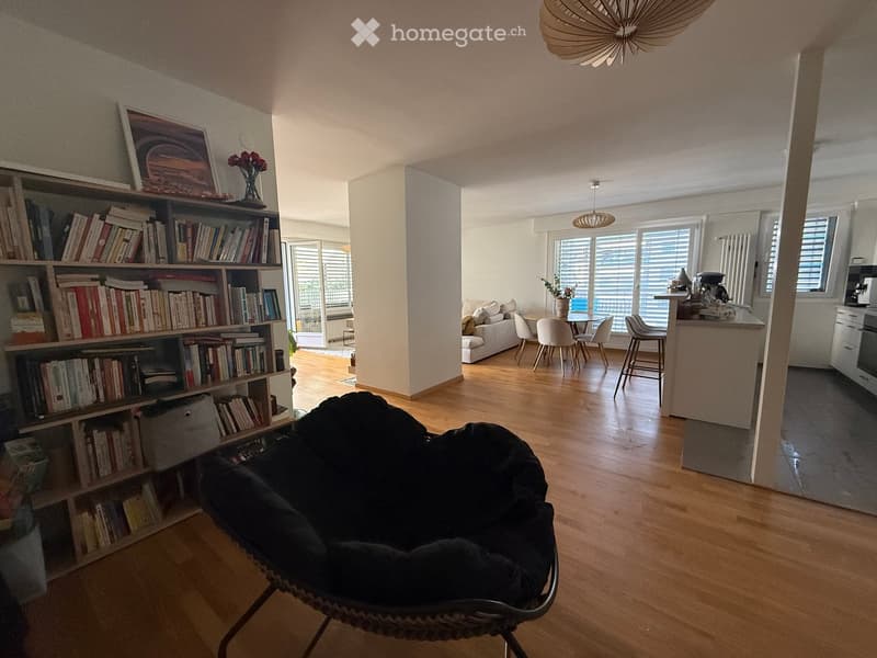 Open-plan living and dining area with wooden flooring, a large bookshelf, a black hanging egg chair, a sofa, a dining table with chairs, and a kitchen area with a counter and appliances.