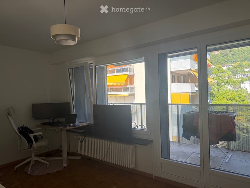 Bright room with large windows and a balcony overlooking a green area. Includes a desk with a laptop, a chair, and a screen on a stand.