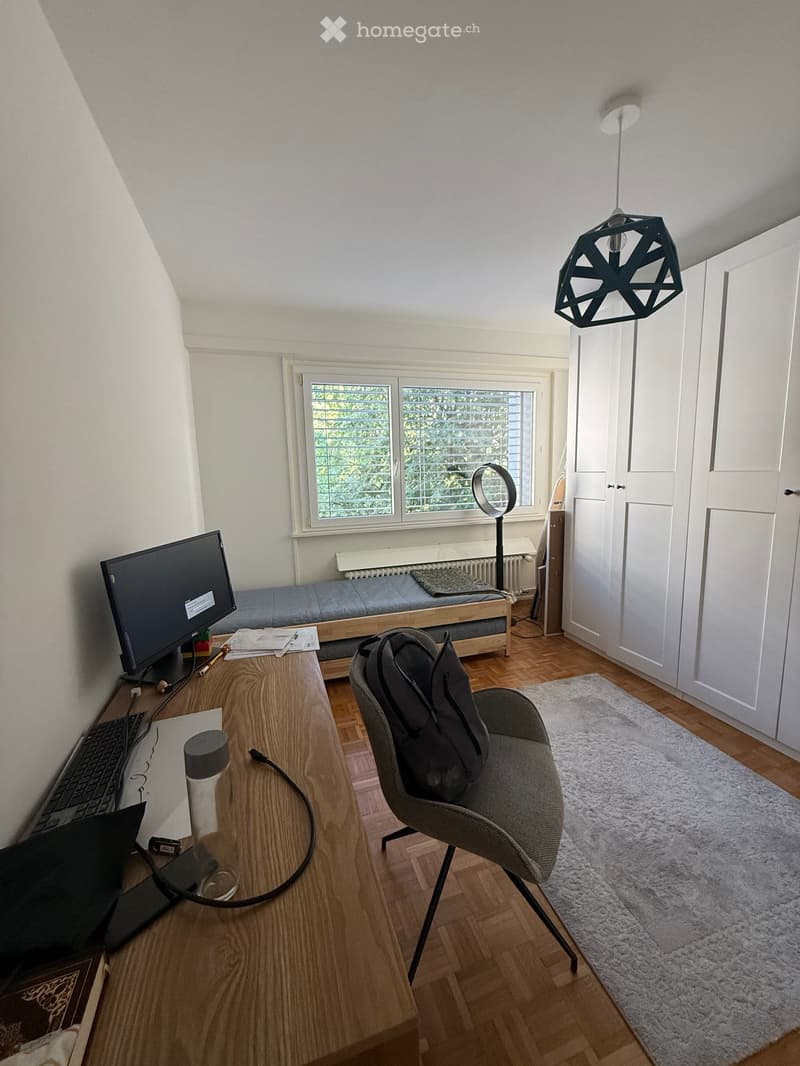A modern, well-lit room with wooden flooring, a large window, a desk with a laptop and keyboard, a comfortable chair with a backpack, a small sofa, and a hanging geometric lamp.