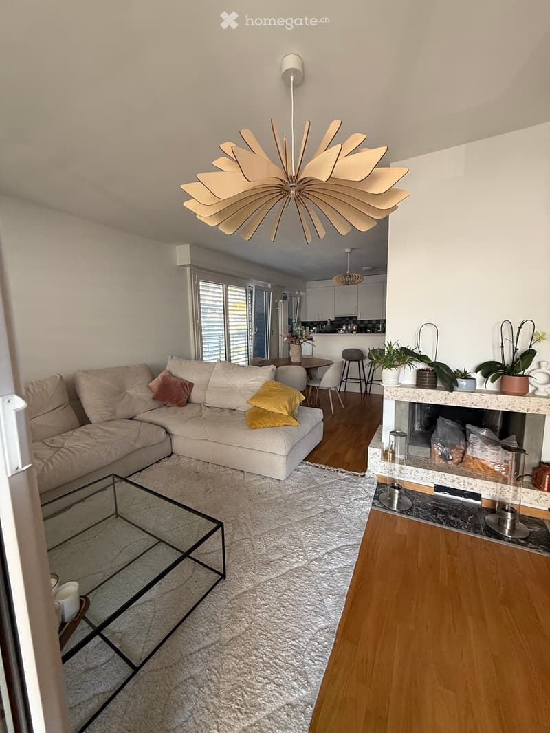 Modern living room with a beige sectional sofa, glass coffee table, fireplace, and dining area visible in the background.