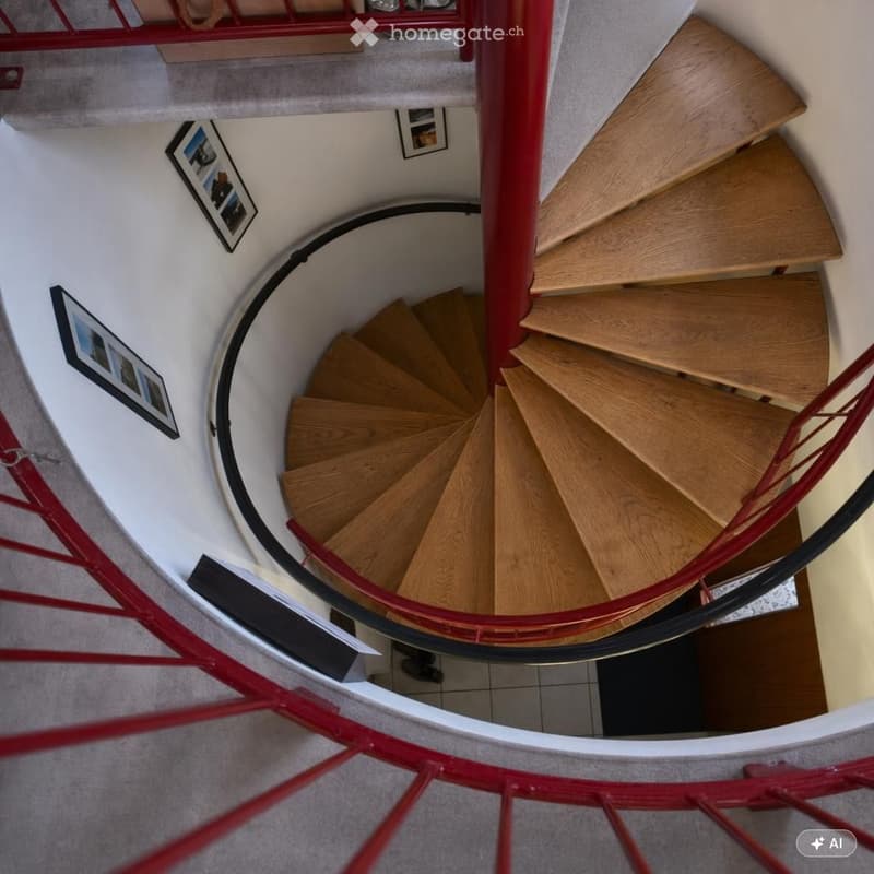 Spiral staircase with wooden steps and red metal railing, surrounded by white walls and framed pictures.