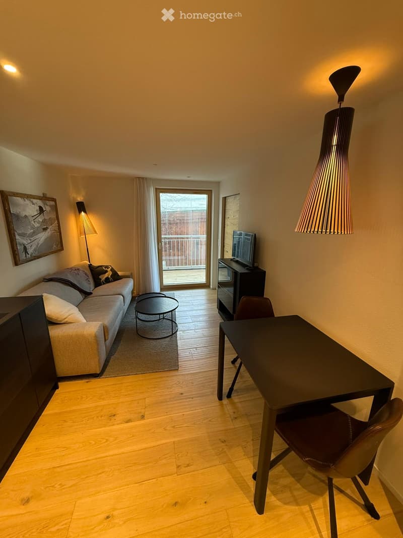 A modern living room with a sofa, coffee table, TV, and a desk area with a chair and table. The room features wooden flooring, warm lighting, and a large window with a view of a balcony.