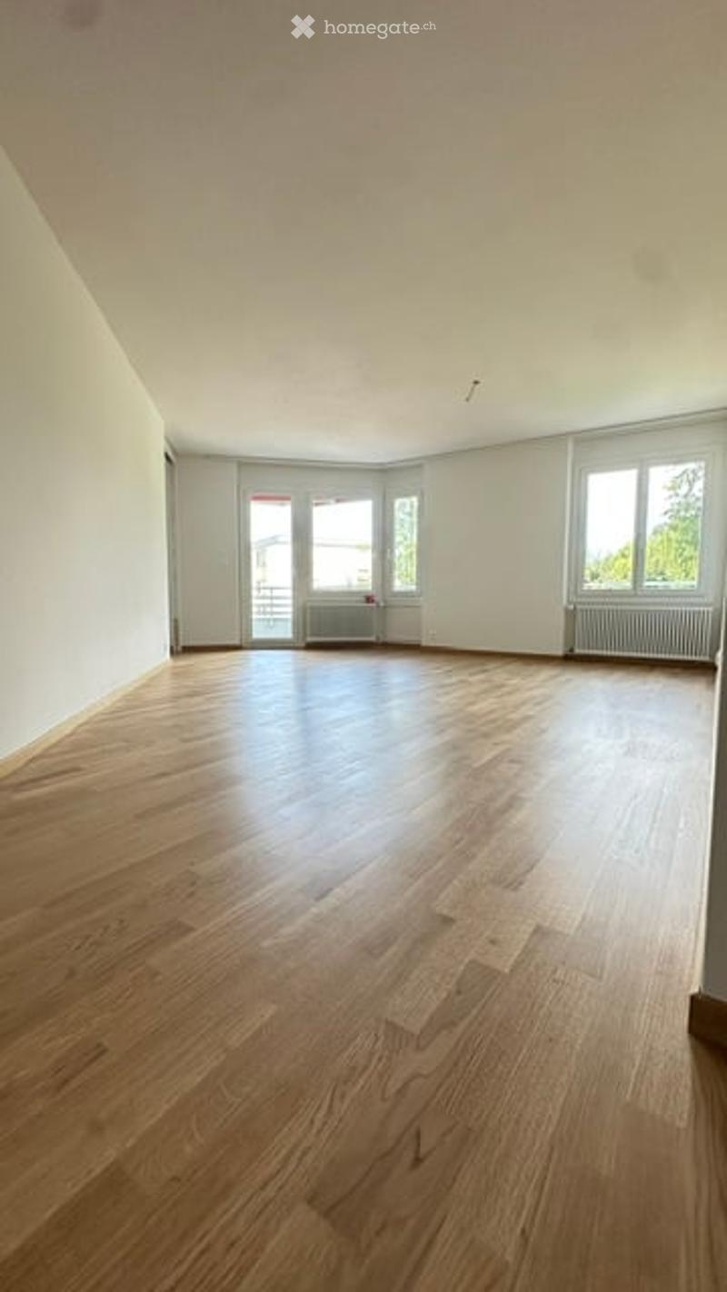 Empty room with light wooden flooring, white walls, and large windows with radiators beneath them.