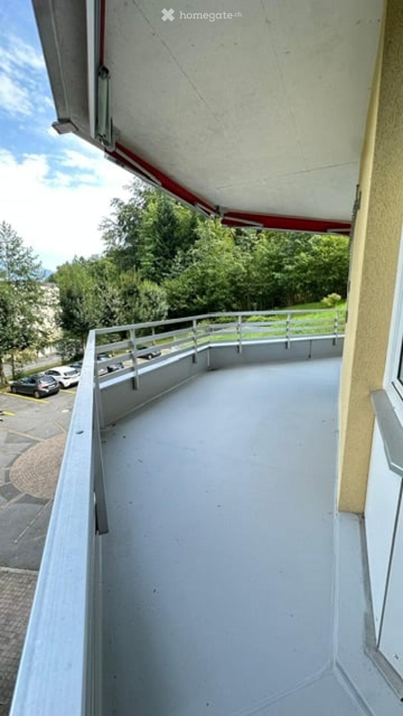 Balcony with white floor, metal railing, and a view of greenery and parked cars.
