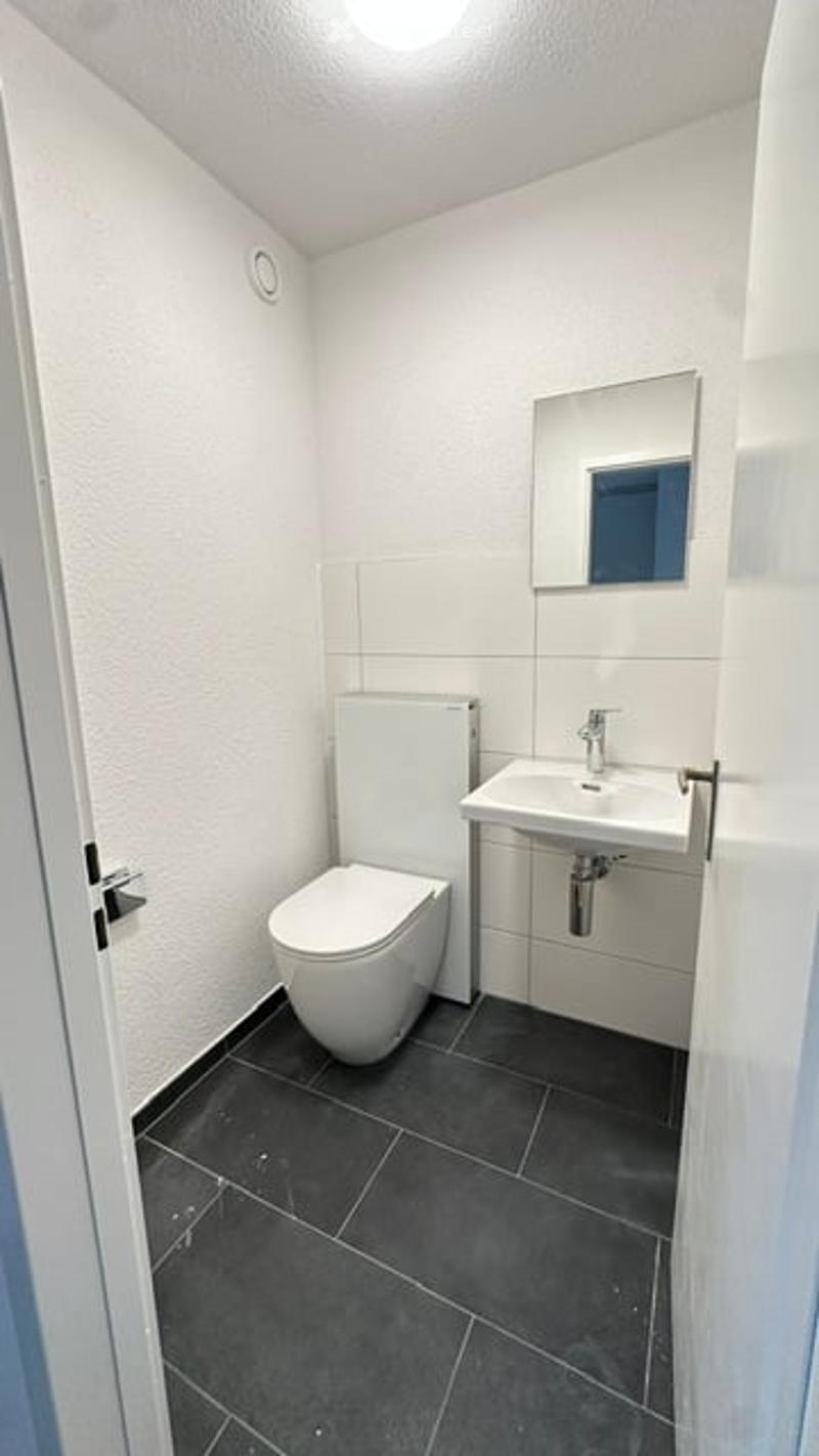 Modern, compact bathroom with white walls, dark tiled floor, toilet, sink, and mirror.