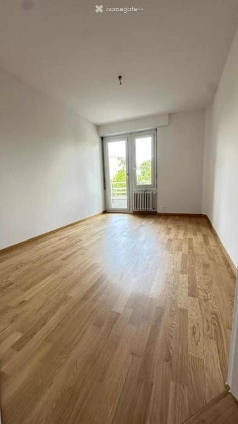 Completely empty room with wooden flooring, white walls, and a large window with a view of greenery outside.