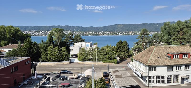 A scenic view from a rooftop terrace overlooking a lake, with surrounding greenery, buildings, and distant hills.