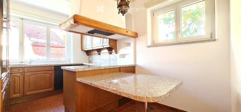 Bright kitchen with wooden cabinets, granite countertop island, sink, and range hood. Large windows provide natural light.