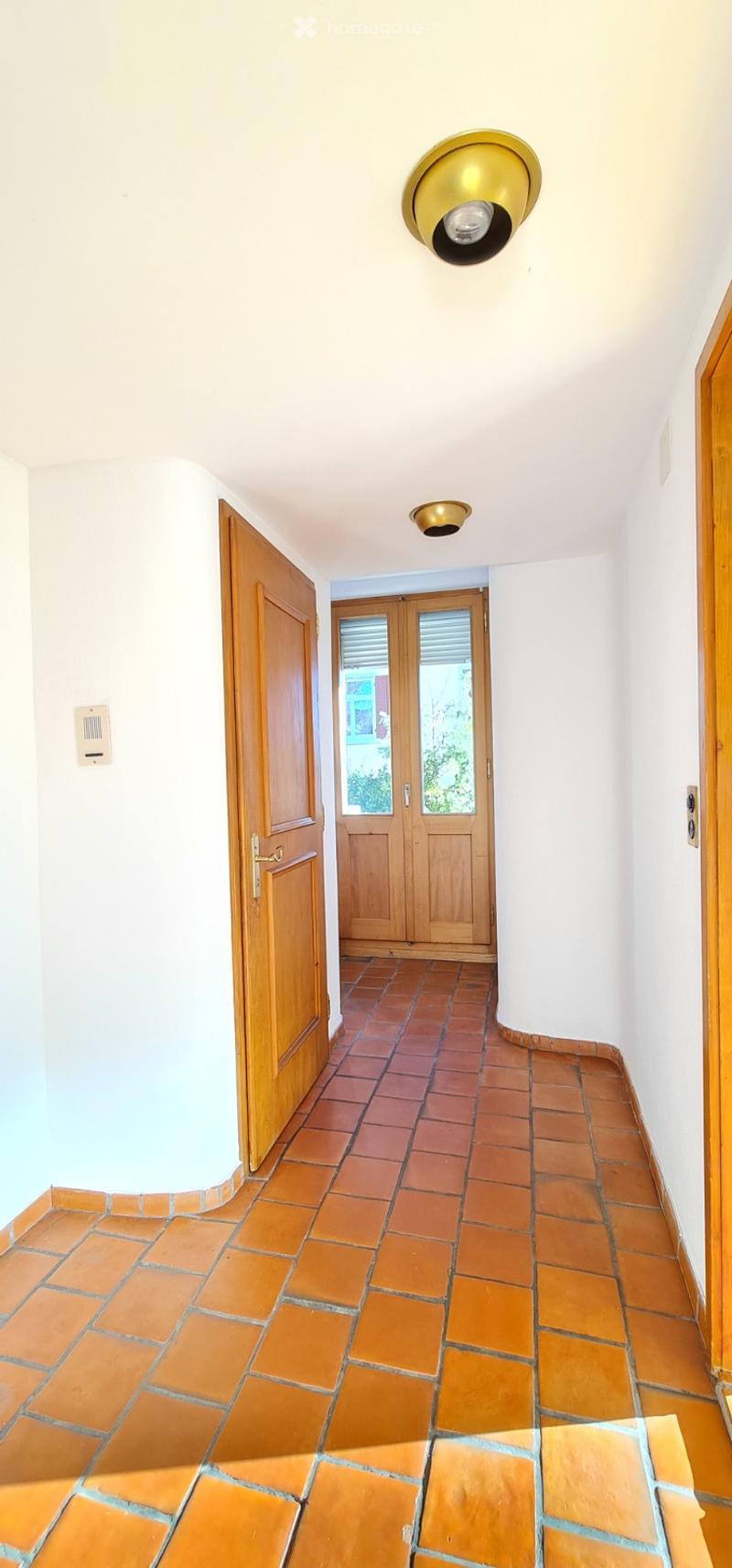 A narrow hallway with terracotta tiled flooring, white walls, and wooden doors. The hallway features a ceiling light fixture and a small window at the far end.