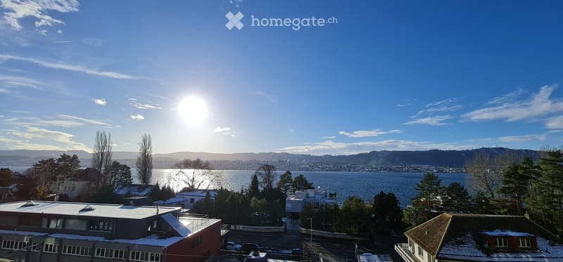 Panoramic view of a cityscape with a lake, mountains, and scattered buildings under a clear blue sky.