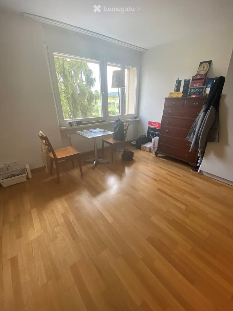 A small, well-lit room with wooden flooring, a window with a view, a table with chairs, a lamp, and a dresser with various items on top.