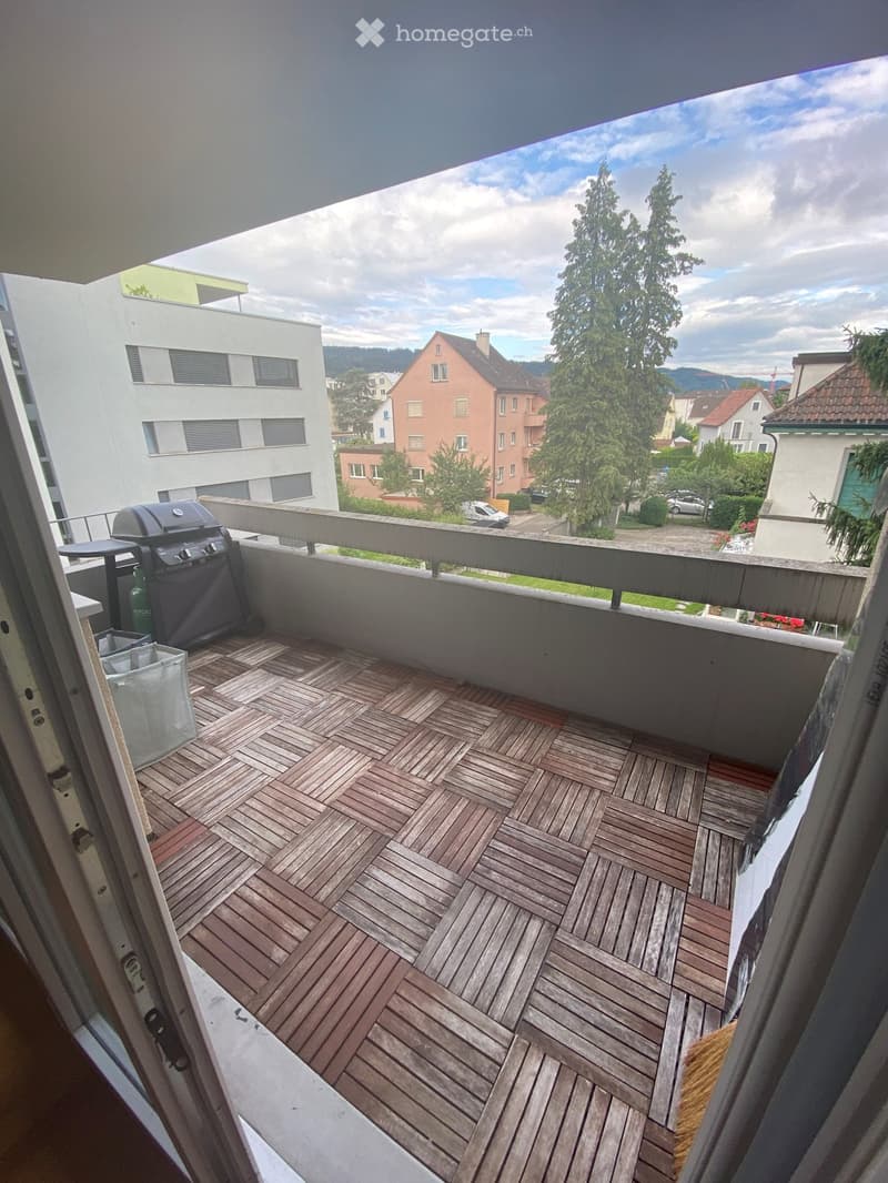Balcony with wooden decking, grill, and table, overlooking a residential area with trees and buildings.