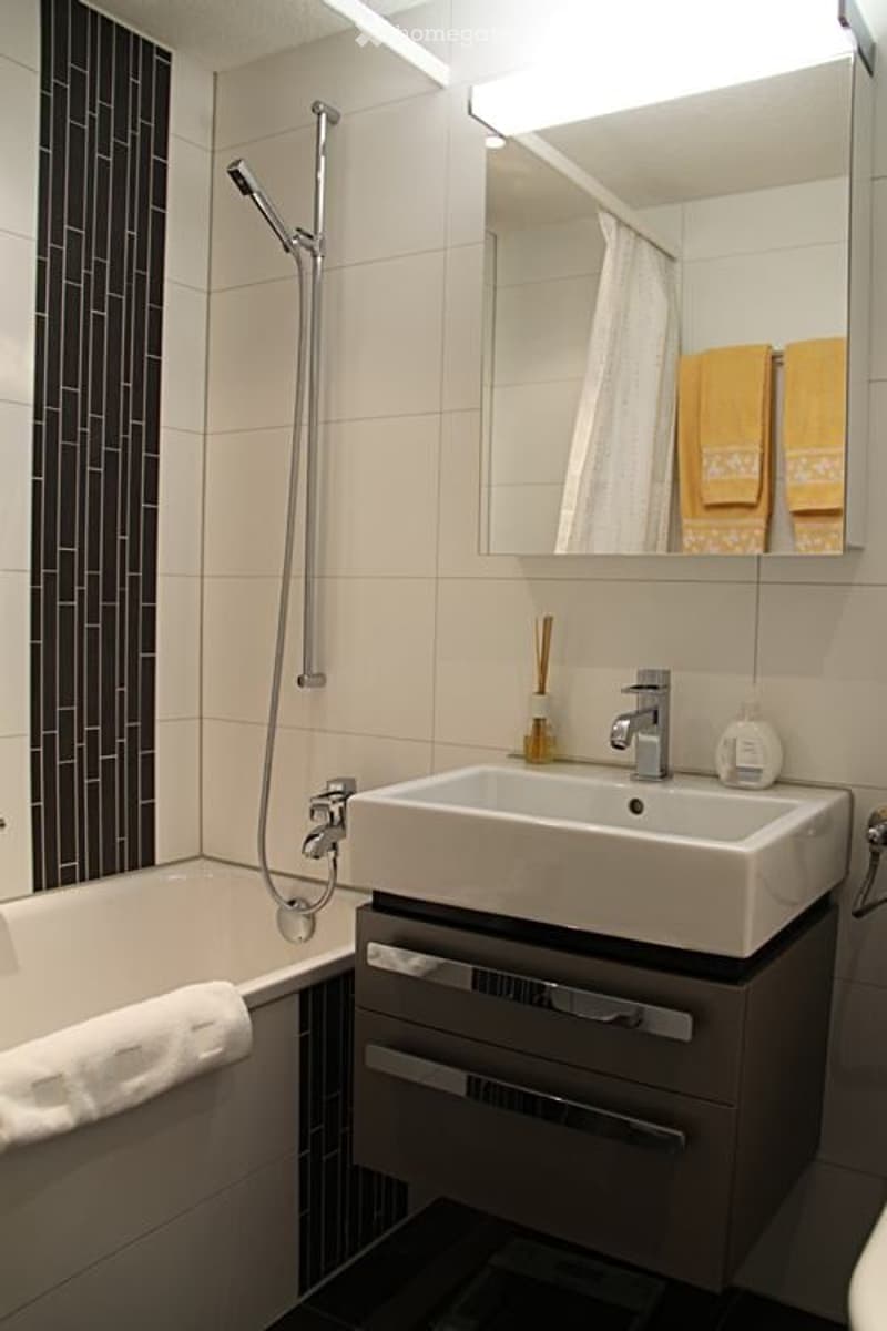 Modern bathroom with a bathtub, sink, mirror, and showerhead. Features include a white sink with a chrome faucet, a built-in vanity with drawers, and a sleek black and white tile design.