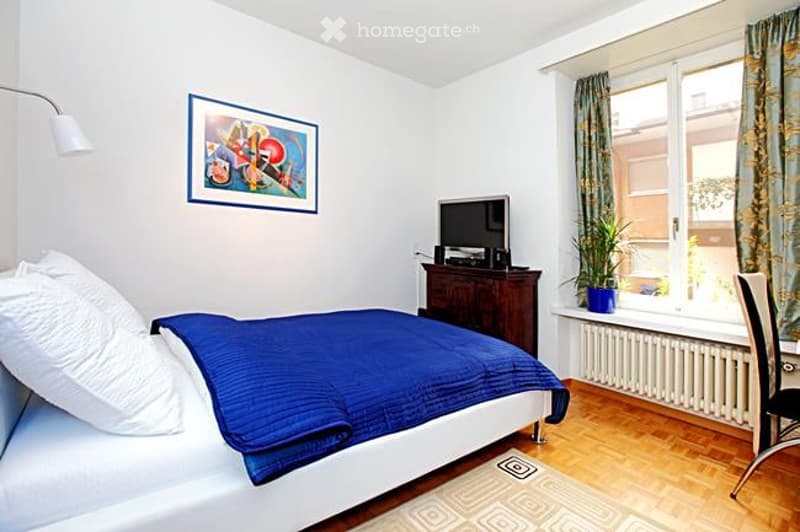 Cozy bedroom with a double bed, nightstand, chair, small table, plant, TV, and a picture frame on the wall.