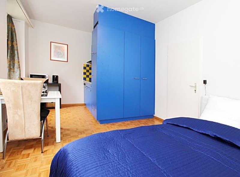 Cozy bedroom with a blue wardrobe, a bed with blue bedding, a small table with a chair, and a framed picture on the wall.
