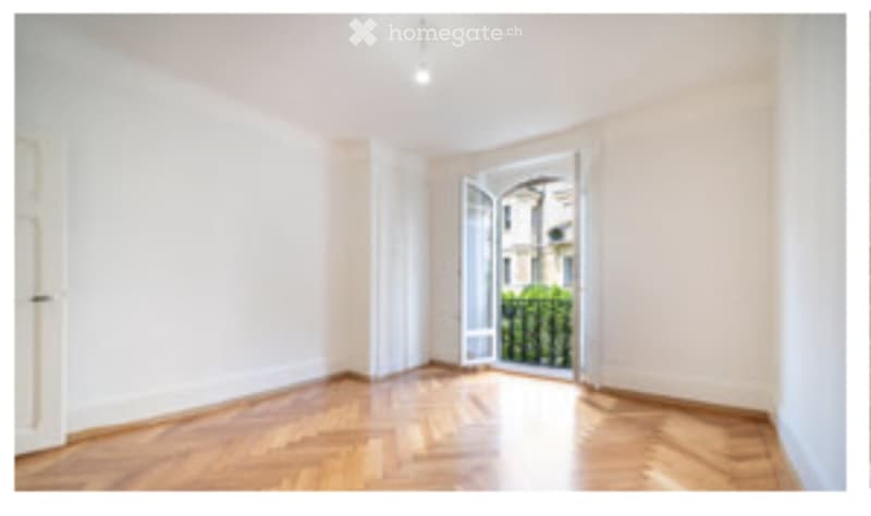Empty room with wooden flooring, white walls, and an open door leading to a balcony with a view of greenery.