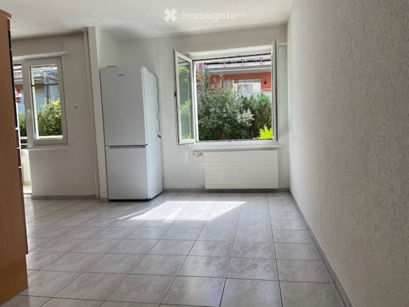 Empty kitchen with tiled floor, white refrigerator, and large windows with open shutters, allowing natural light to fill the space.