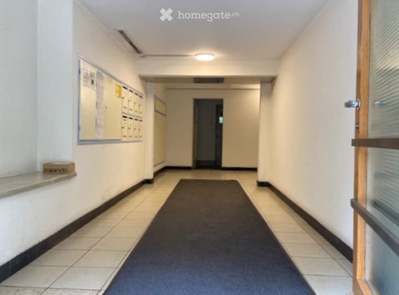 Clean, well-lit hallway with tiled flooring, a dark runner, and a bulletin board on one wall.