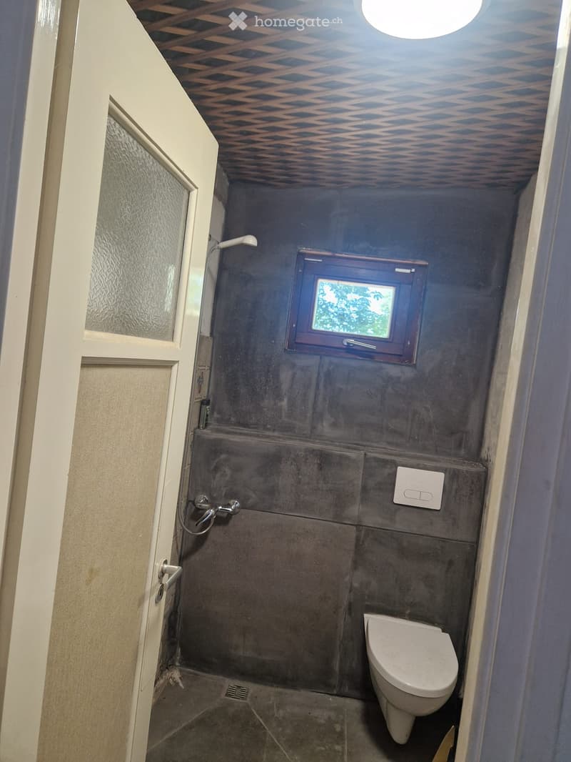 Small bathroom with dark grey tiled walls and floor, a toilet, a shower area with a handheld showerhead, and a small window above the toilet.