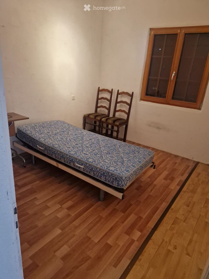A small, furnished room with wooden flooring, a single bed with a blue patterned mattress, two wooden chairs, and a small table.