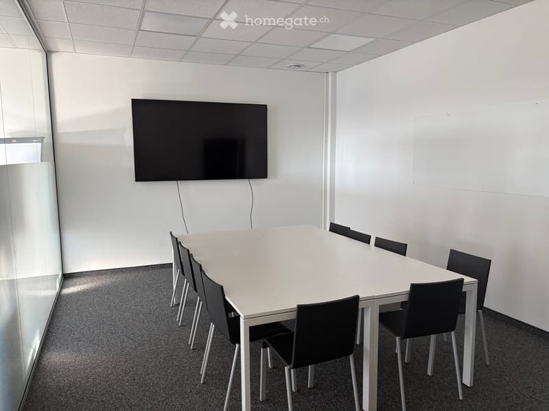 Empty meeting room with a long white table, black chairs, and a mounted flat screen TV on the wall.