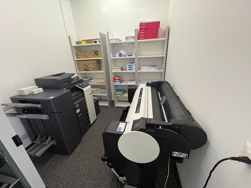 Office room with two large printers, shelves with books and boxes, and a carpeted floor.