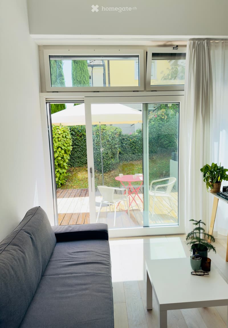 Modern living room with a grey sofa, white coffee table, and large windows leading to a balcony with outdoor furniture.
