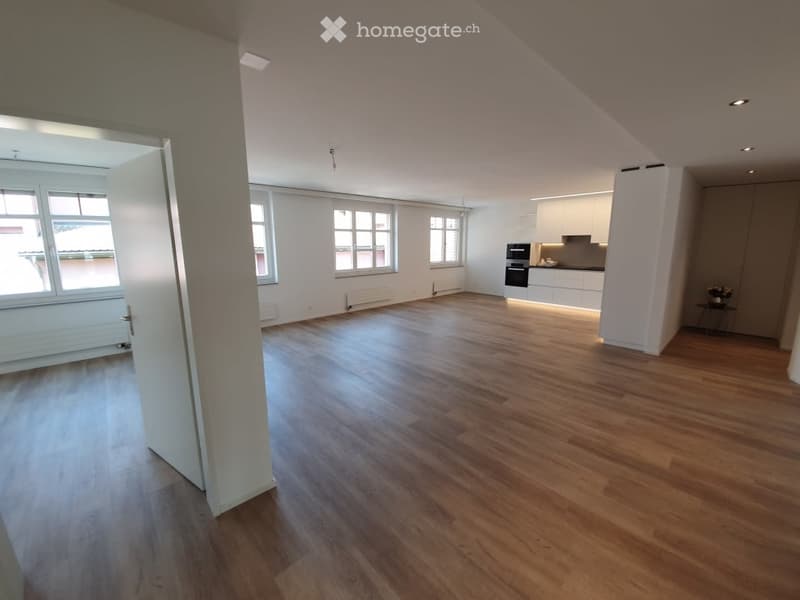 Bright, empty room with wooden flooring, white walls, large windows, and a small kitchen area visible in the background.