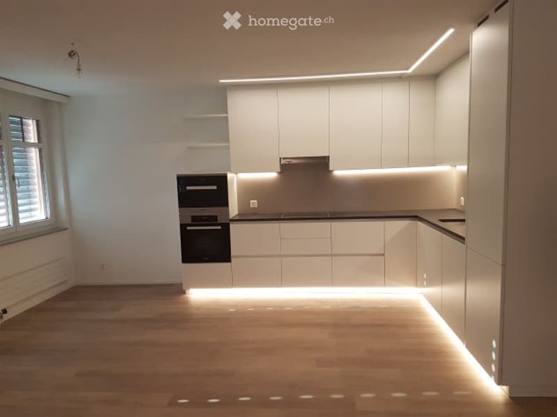 Modern kitchen with white cabinets, black oven, and sleek countertop, illuminated by integrated LED lighting under the cabinets and on the floor.