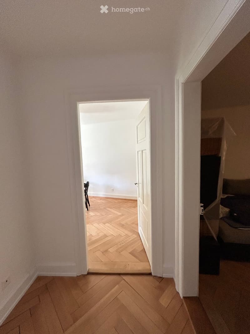 A narrow hallway with light pink walls and wooden flooring, leading to an open doorway that reveals another room with a visible chair.