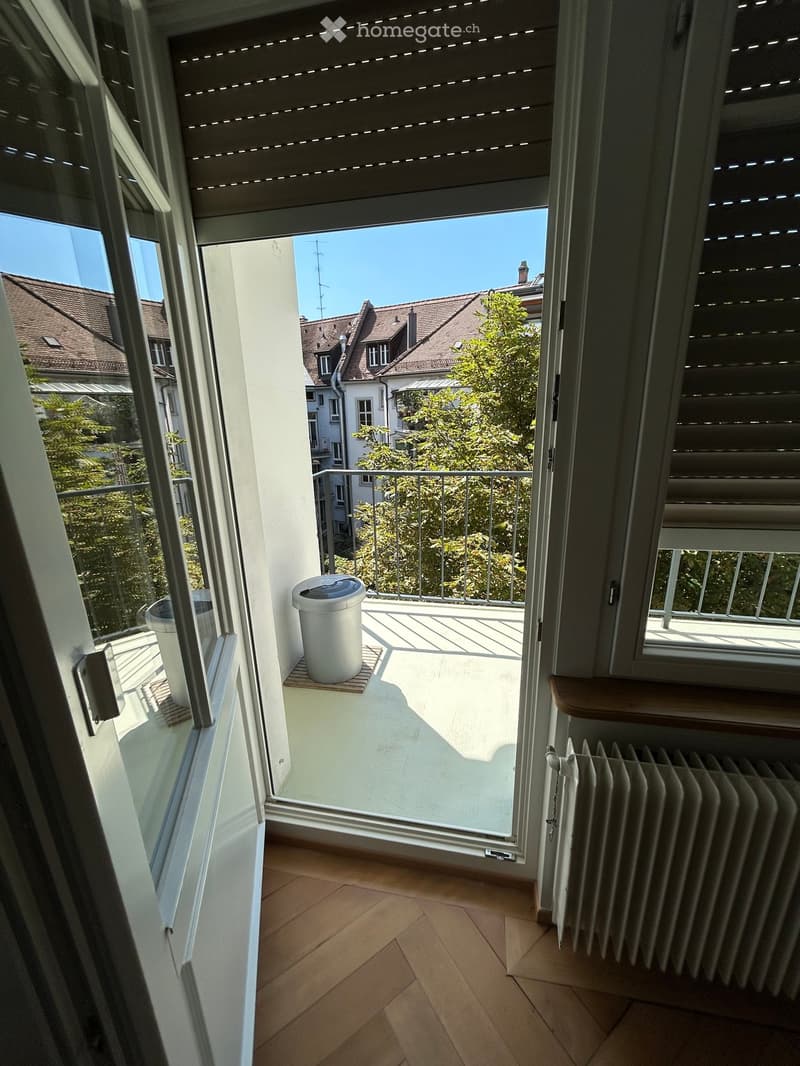 Balcony with wooden floor, white trash bin, metal railing, and view of neighboring houses and trees.