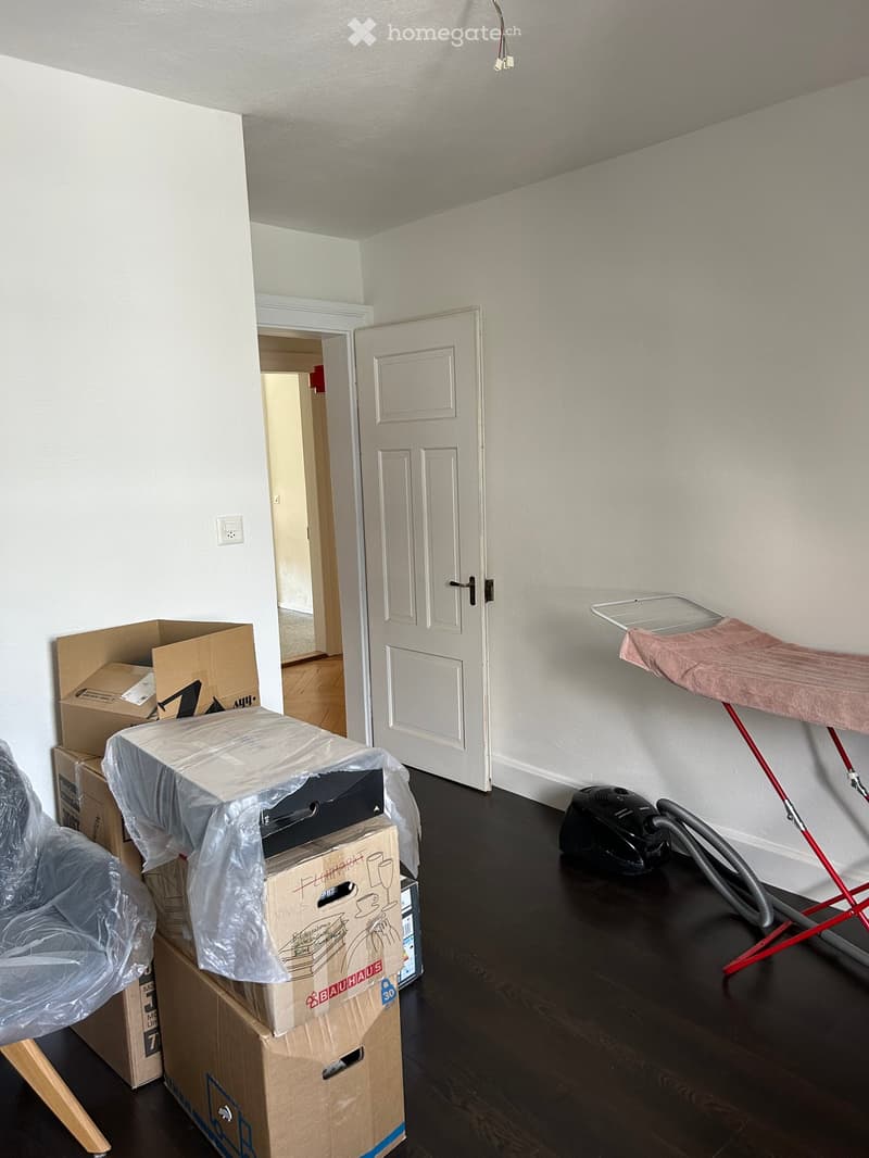 Empty room with dark wooden flooring, white walls, and several cardboard boxes stacked near the door. A vacuum cleaner and an ironing board are also visible.