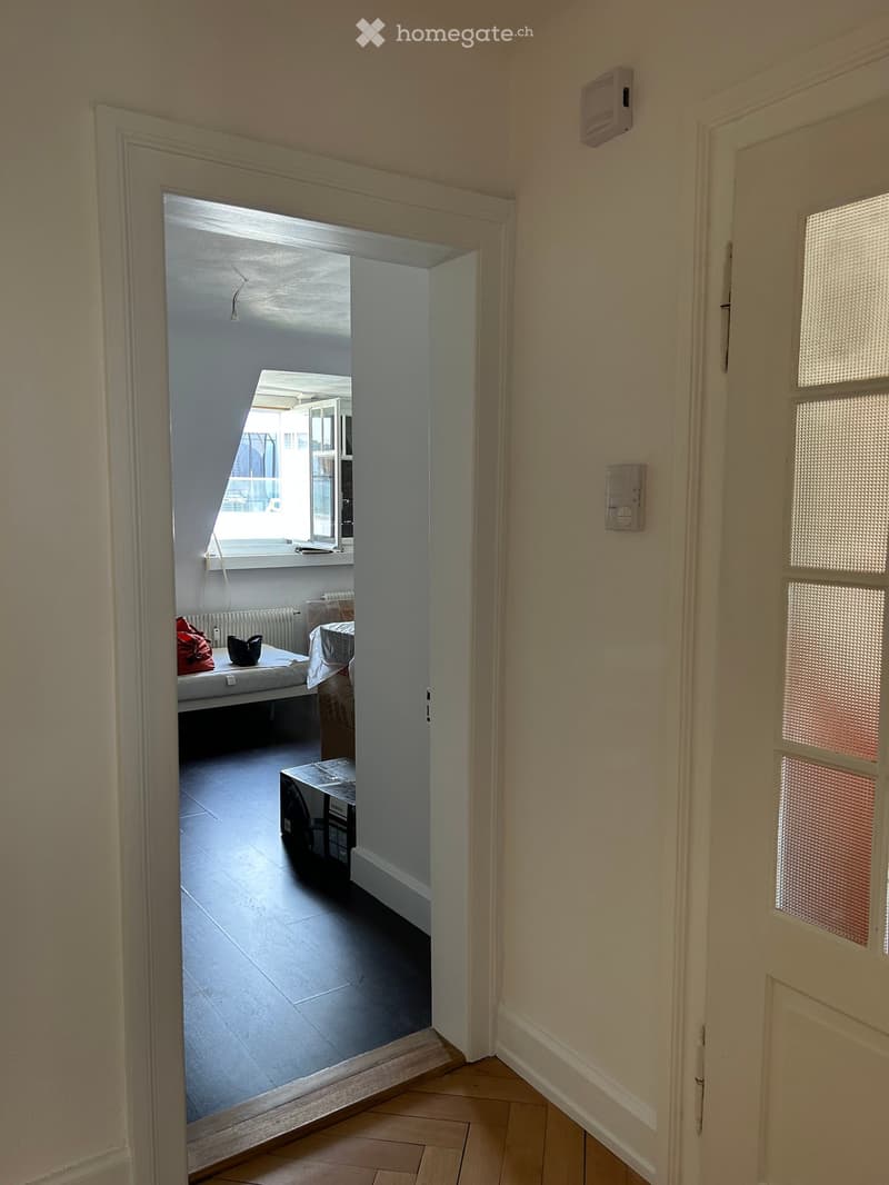 A narrow hallway with light-colored walls and wooden flooring, leading to a room with a bed, table, and window.