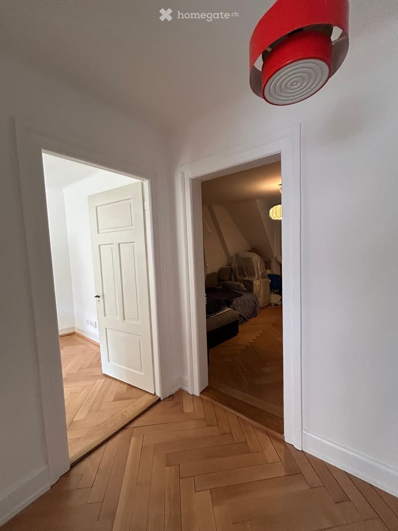 Hallway with wooden flooring, white walls, and two open doors leading to other rooms. A red decorative light fixture is mounted on the ceiling.