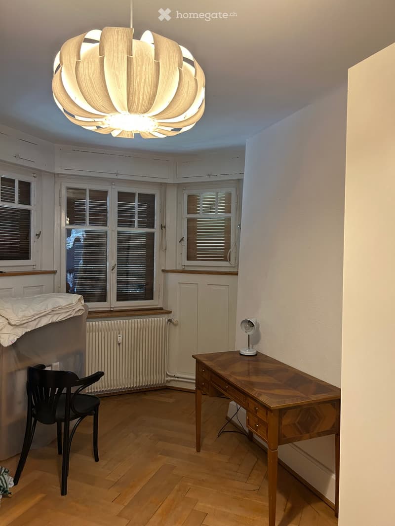 A furnished room with wooden flooring, a large window with blinds, a modern chandelier, a wooden desk, a black chair, and a small table lamp.