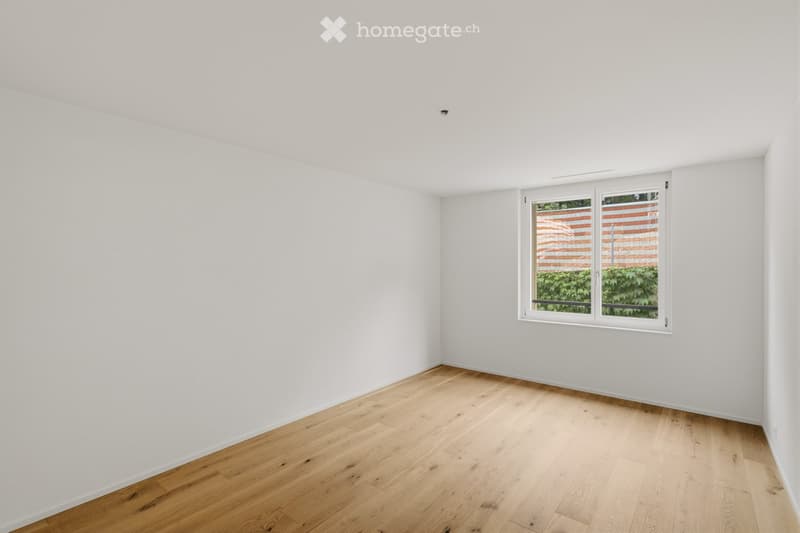Empty room with white walls and light wooden flooring, featuring a large window with a view of greenery outside.