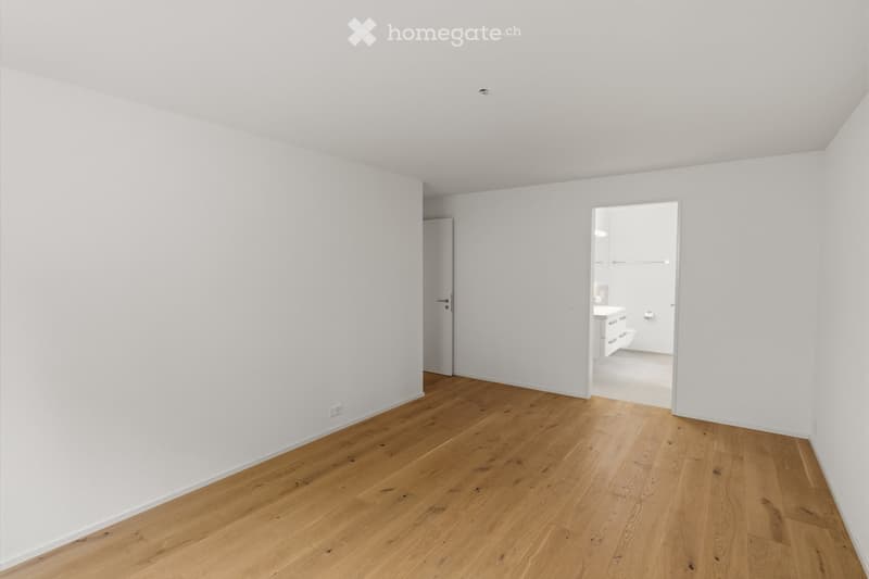 Empty room with light wooden flooring and white walls, featuring a doorway leading to a small bathroom with white cabinets and a sink.