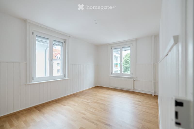 Completely empty room with bare white walls, wooden floor, and two windows with white frames. No furniture or equipment.
