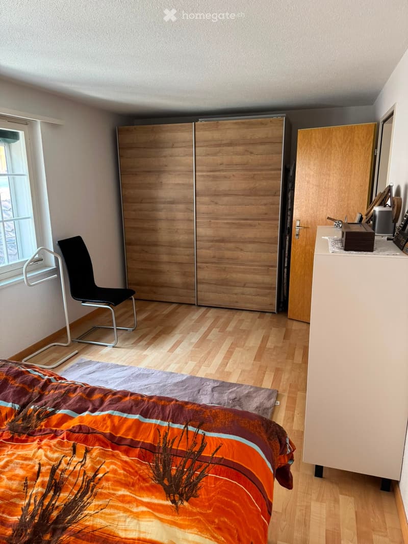 A modern bedroom with a wooden floor, a large bed with an orange and brown blanket, a black chair, a large wooden wardrobe, and a small table with a mirror.