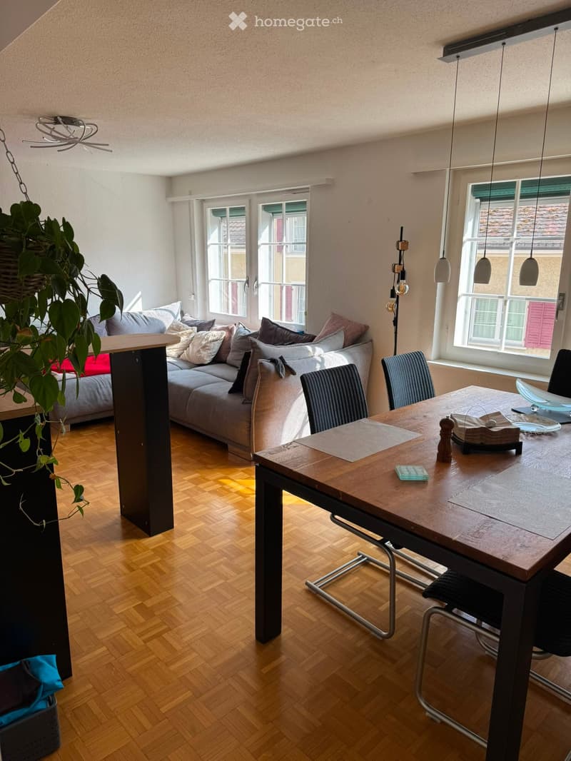 A modern living room with a grey sectional sofa, wooden dining table, chairs, pendant lights, and a hanging plant. The room features large windows and a wooden floor.