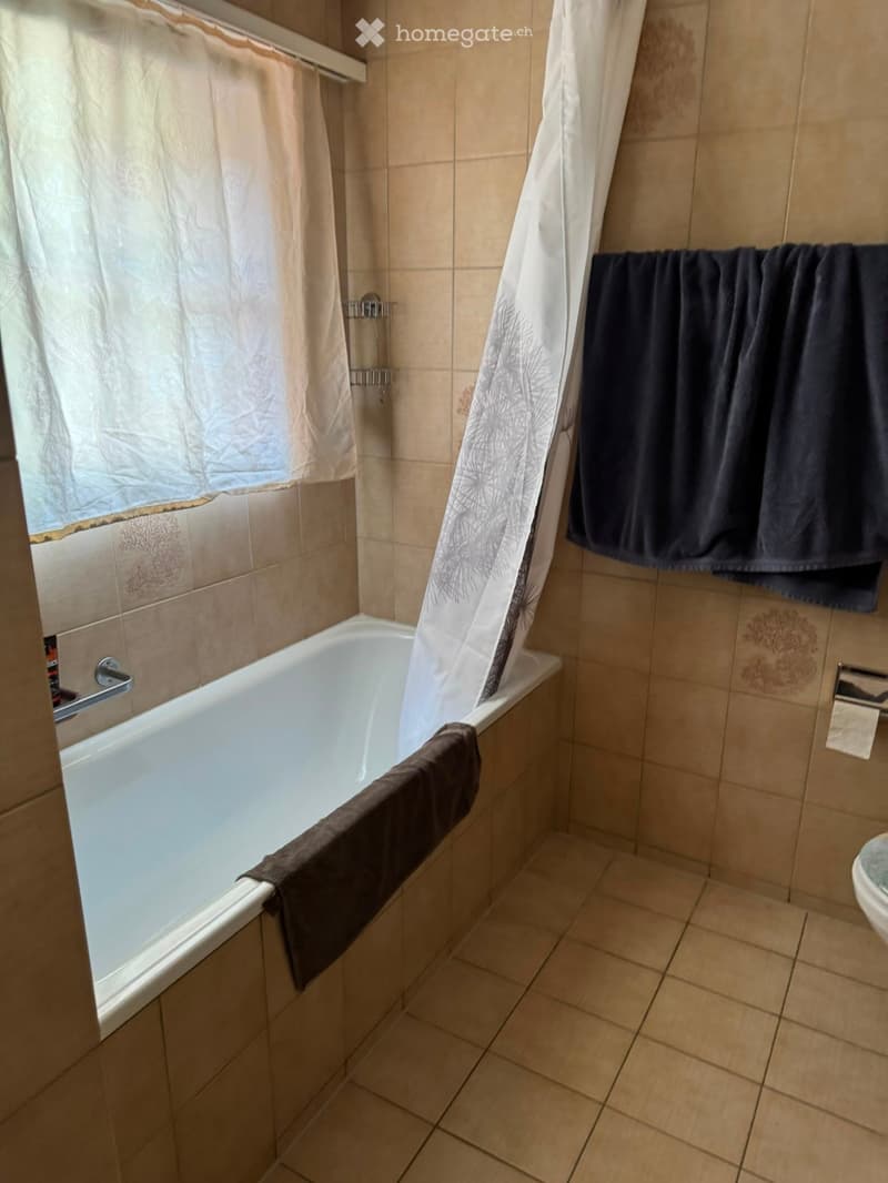 Bathroom with a bathtub, shower curtain, towels, and a toilet. Beige tiled walls and floor.