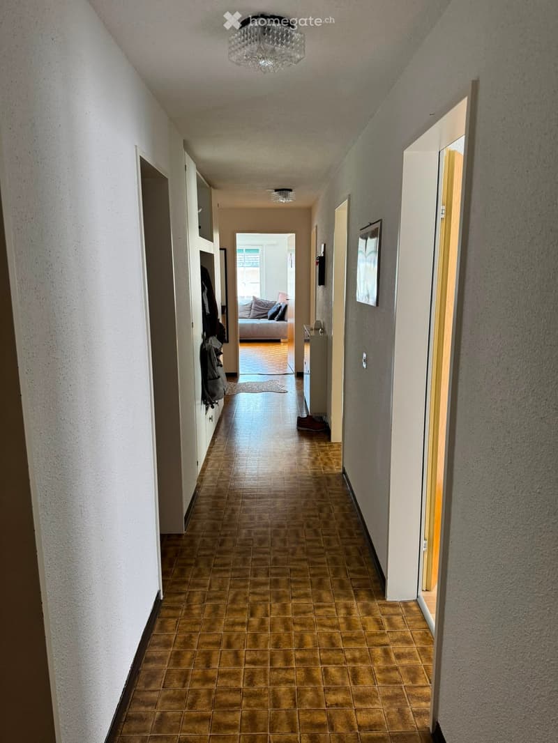 A long, narrow hallway with tiled flooring, white walls, and several doors leading to different rooms. A chandelier hangs from the ceiling, and a glimpse of a living area with a sofa and pillows is visible at the end of the hallway.