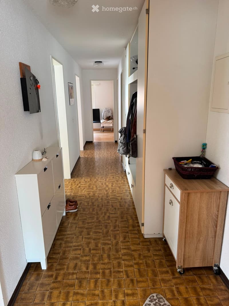 A long hallway with tiled flooring, white walls, and several doors leading to different rooms. There are small cabinets and a coat rack with jackets and shoes visible.