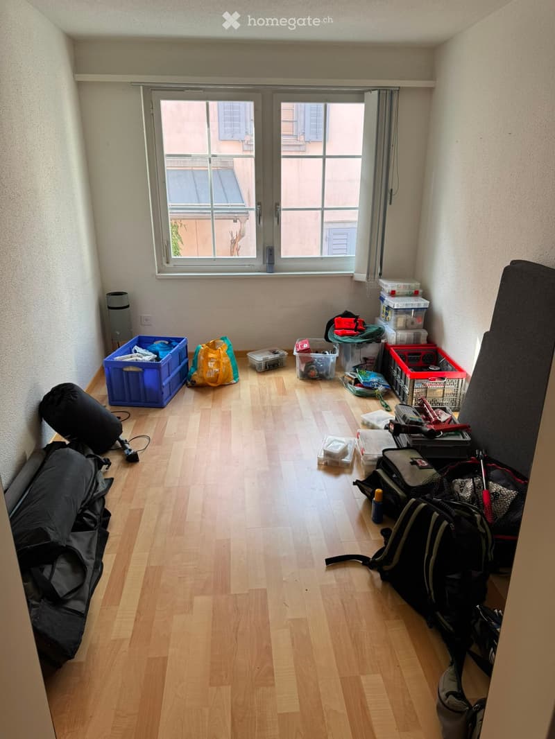 Empty room with wooden flooring, light-colored walls, and a large window with a view of a neighboring building. Various storage boxes and bags are scattered around the room.