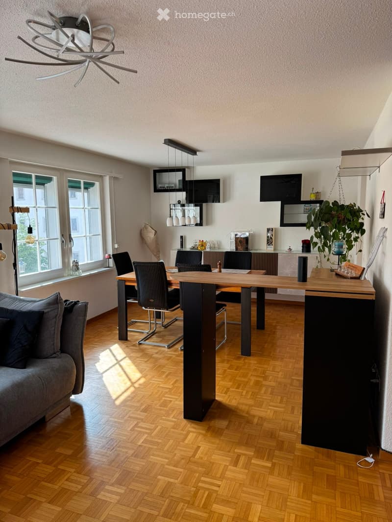 Open-plan living and dining area with wooden flooring, grey sofa, dining table with chairs, kitchen island, pendant lights, wall-mounted TV, and a large plant near the window.
