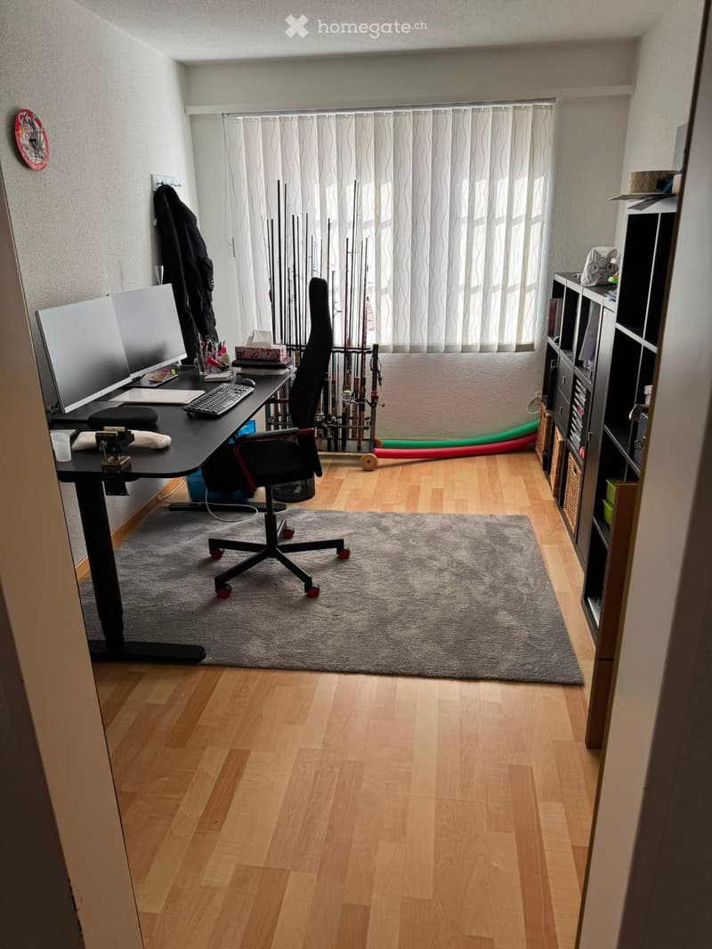 A well-lit room with wooden flooring, a desk setup including a monitor, keyboard, and chair, a shelf with various items, and a curtained window.
