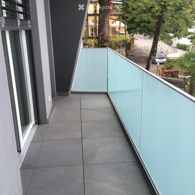 Modern balcony with grey tiled floor and glass railing, overlooking a street with trees, outdoor seating, and a parked car.