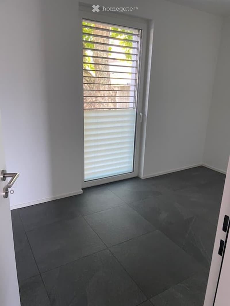 Empty room with dark grey tiled floor, white walls, and a large sliding glass door with blinds leading to an outdoor area.