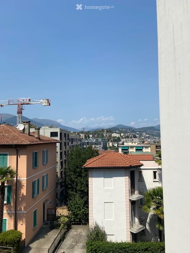 View of a cityscape with multiple buildings, including residential structures and a construction crane in the background. The scene includes a mix of architectural styles and greenery.